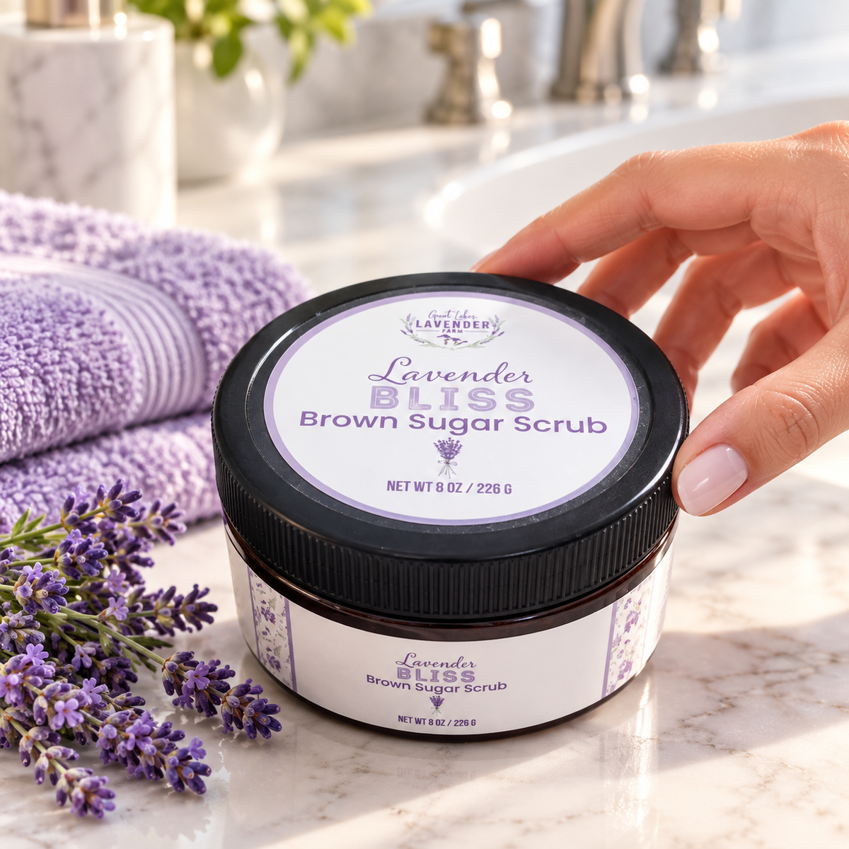 Hand reaching for a jar of Lavender Bliss Brown Sugar Scrub on a marble counter with purple towels and lavender