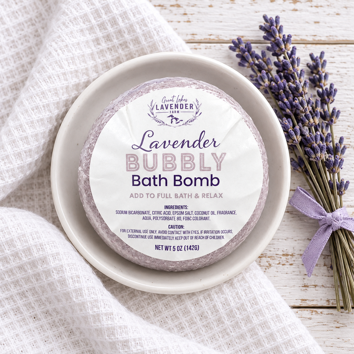 Lavender Bubbly Bath Bomb in label wrap on a white dish with a tied lavender bundle on a towel