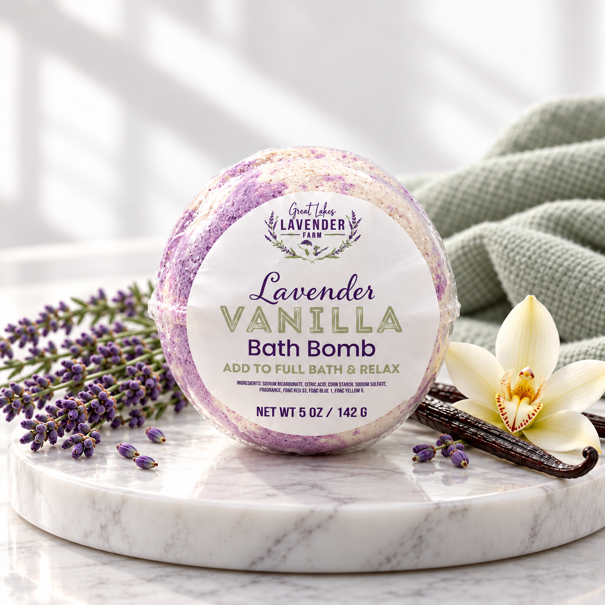 Lavender Vanilla Bath Bomb, shrink-wrapped, on a marble tray with lavender, vanilla beans, a vanilla orchid and green towel