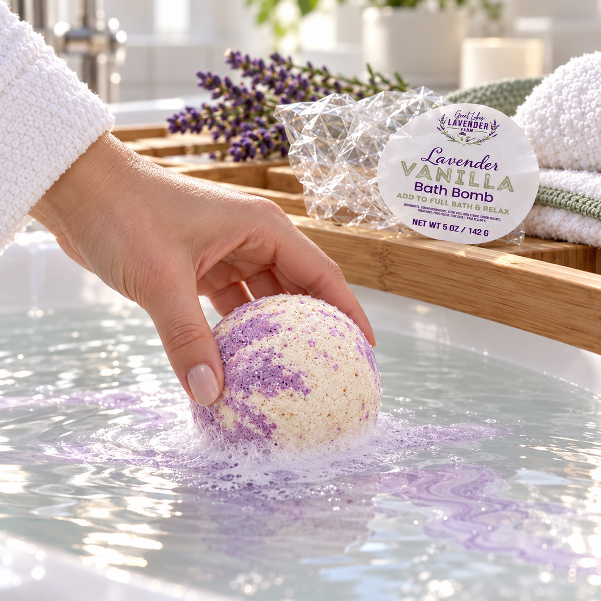 Hand lowering a purple and cream Lavender Vanilla Bath Bomb into bathwater, wrapped bath bomb on a tray behind