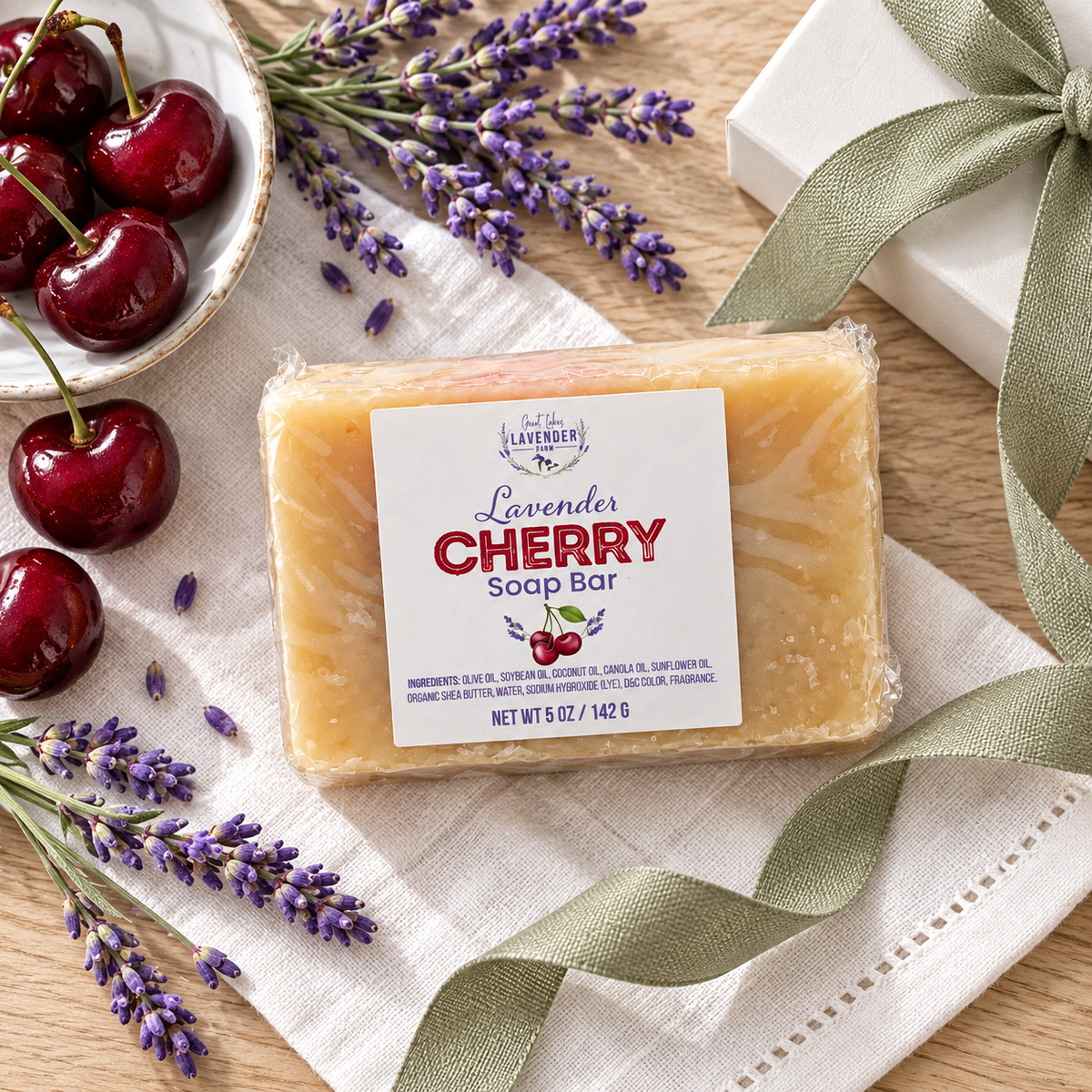 Lavender Cherry Soap Bar on a linen napkin with a bowl of cherries, lavender and a gift box with green ribbon