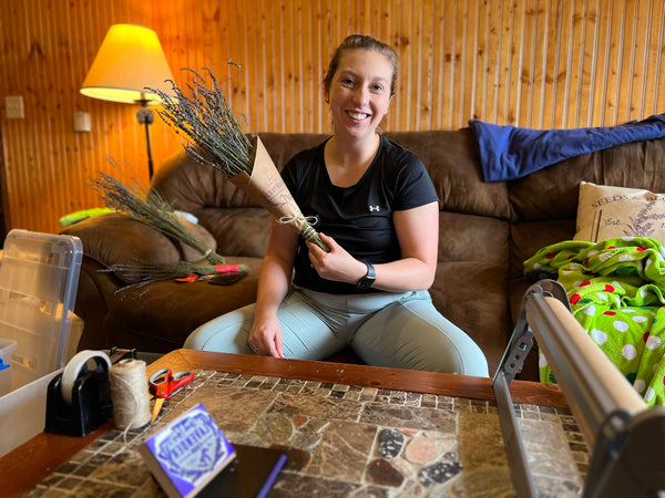 Smiling woman holding a kraft-wrapped dried lavender bouquet at a table of twine and wrapping paper