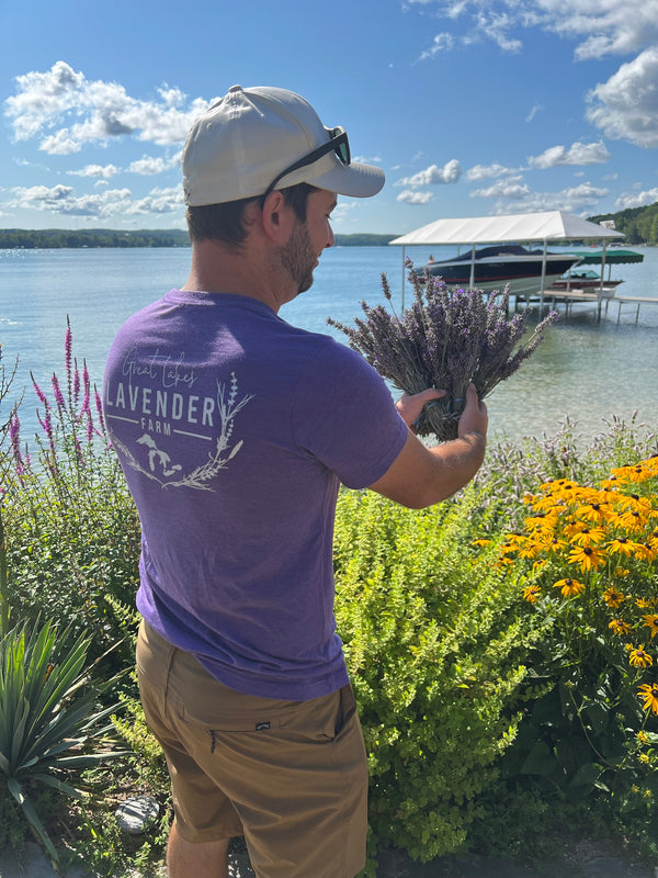 Man in a Great Lakes Lavender Farm shirt holding a lavender bundle beside a lake
