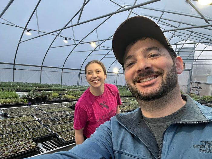 Selfie of two smiling people inside the greenhouse full of seedling trays